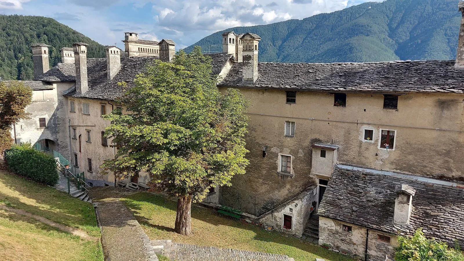 Case in pietra del centro storico di Craveggia con tetti in piode e alti comignoli, un ippocastano nel prato e montagne boscose sullo sfondo