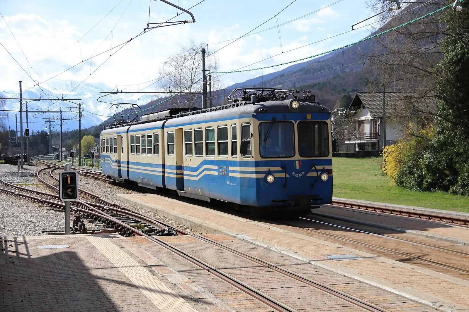 Elettrotreno bianco e azzurro della SSIF in arrivo sui binari a scartamento metrico, con montagne innevate sullo sfondo