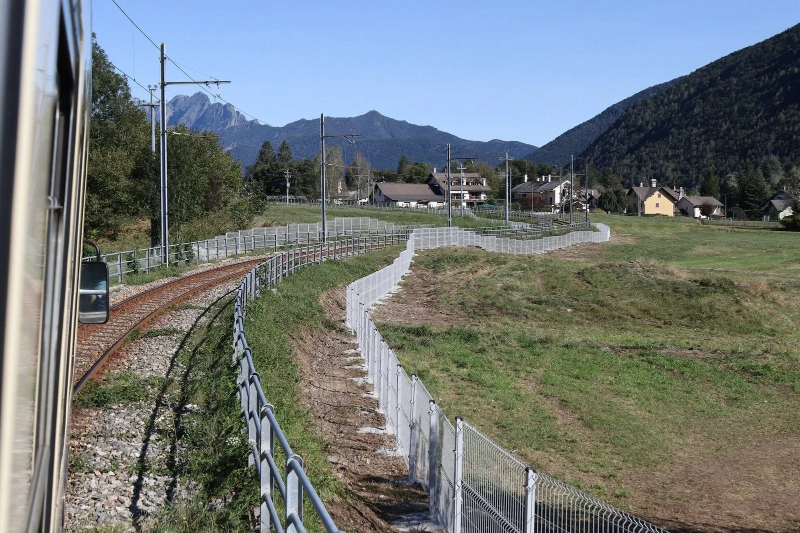 Il binario della Ferrovia Vigezzina visto dal finestrino del treno tra Buttogno e Santa Maria Maggiore, con prati, case e montagne
