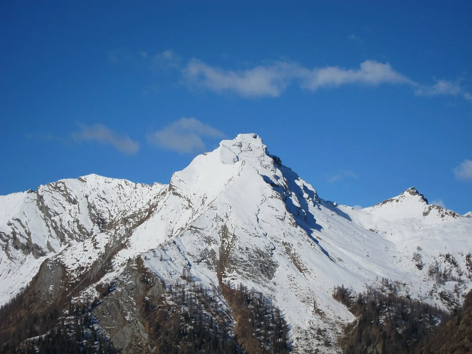 La Pioda di Crana innevata sotto un cielo blu, vista in inverno dalla Piana di Vigezzo, con boschi di larici alla base