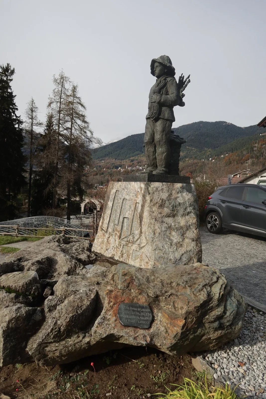 La statua in bronzo del piccolo spazzacamino a Malesco, su un basamento di pietra, con le montagne della Valle Vigezzo in autunno